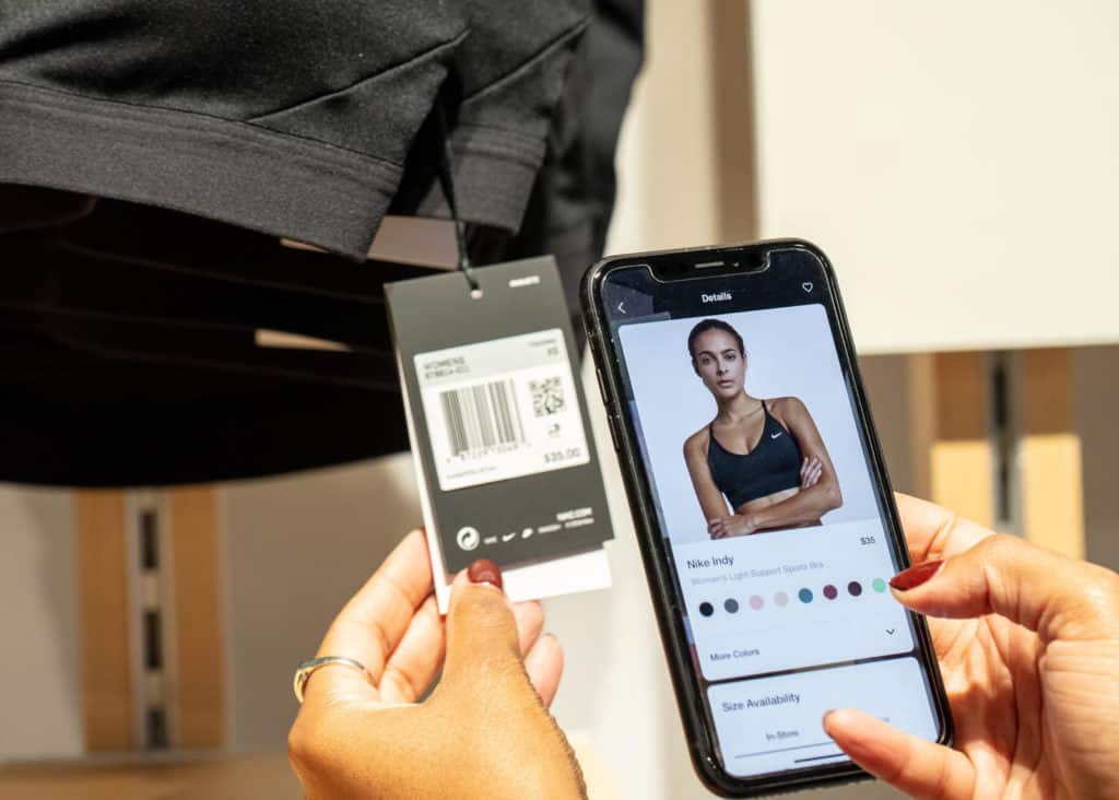 Woman's hands using the Nike Scan feature via the Nike App at Retail to scan a QR code on an in store sports bra. The smartphone photo displays the product page at Nike's ecommerce site.