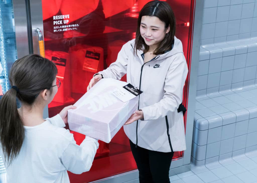 Woman taking a Nike shoe box from a female sales associate using the reserve and pick up in store feature at the Nike Tokyo store.